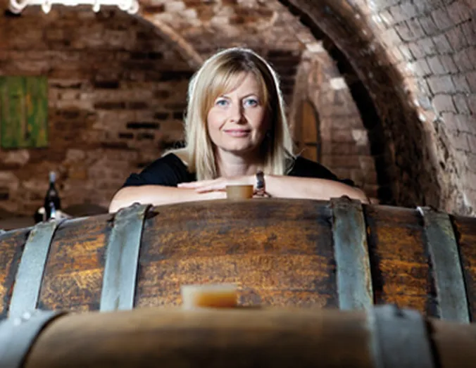 Woman with blonde hair smiling behind large wooden barrels in a rustic winery cellar.