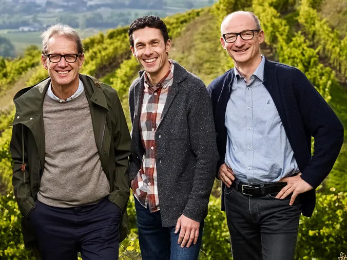 Three men smiling and posing together on a sunny day in a vineyard, with green rows of grapevines in the background.