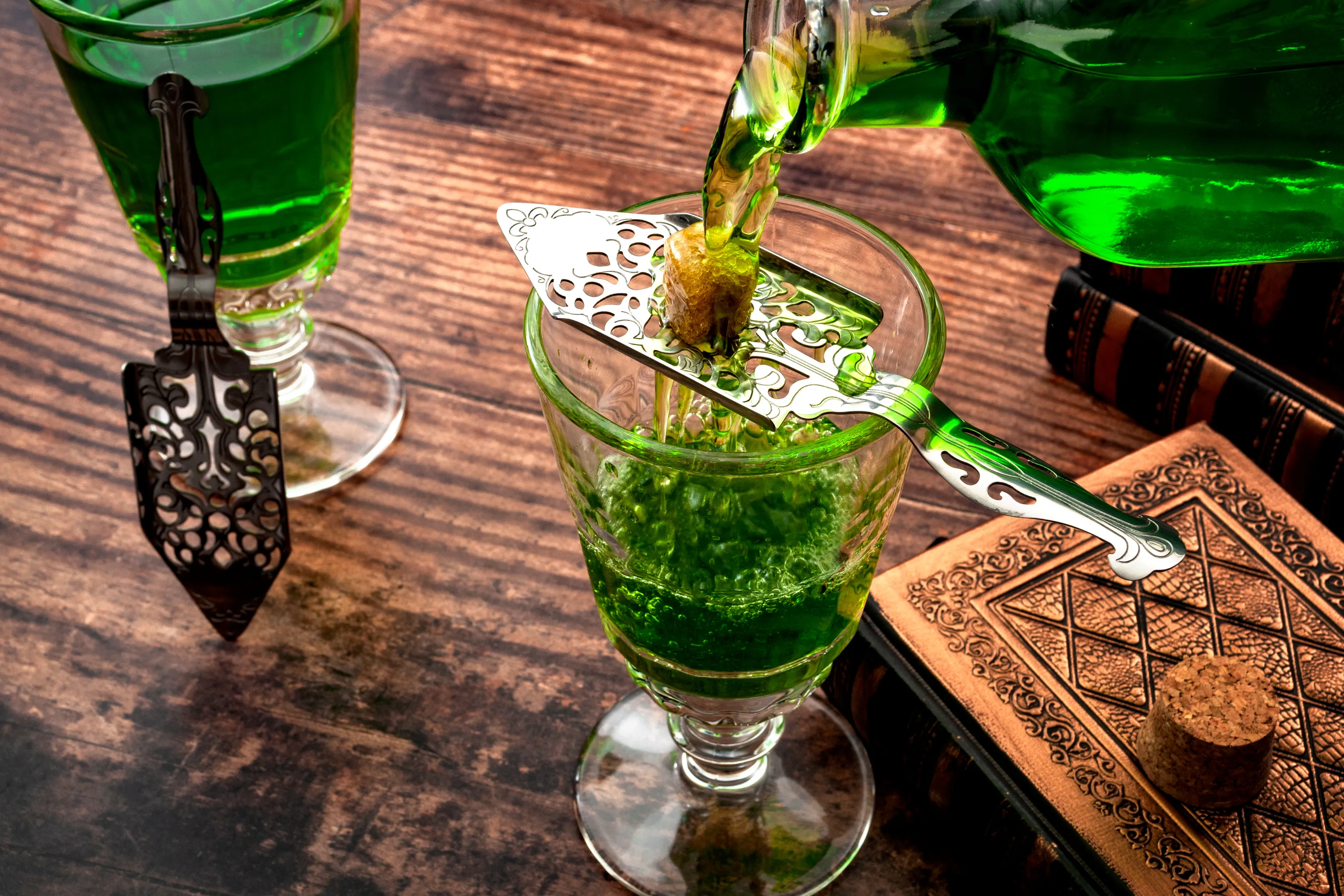 Absinth wird durch einen gelochten Löffel mit einem Zuckerwürfel in ein Glas gegossen, das auf einem rustikalen Tisch neben einem Korken und einem geprägten Buch steht.