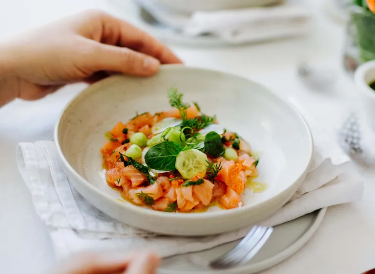 Eine Hand, die eine weiße Schale mit frischem Saiblings-Ceviche hält, garniert mit Gurke, Kräutern und grünen Erbsen, auf einem gedeckten Tisch.