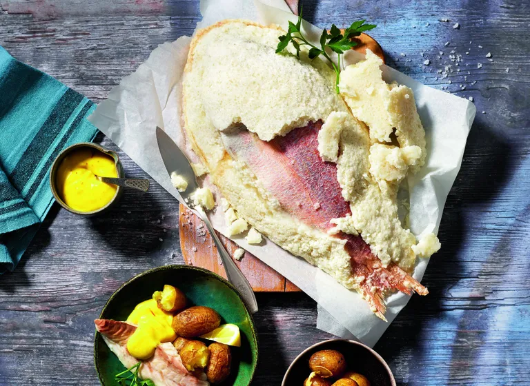 Salt-crusted fish with herbs on a wooden board, served with potatoes, lemon, and yellow sauce; teal napkin on the side.