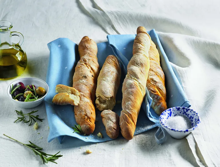 Drei Baguettes auf einem blauen Tischtuch mit Olivenöl, eine Schale mit gewürzten Oliven, Rosmarin und Salz auf einer weißen Tischdecke.