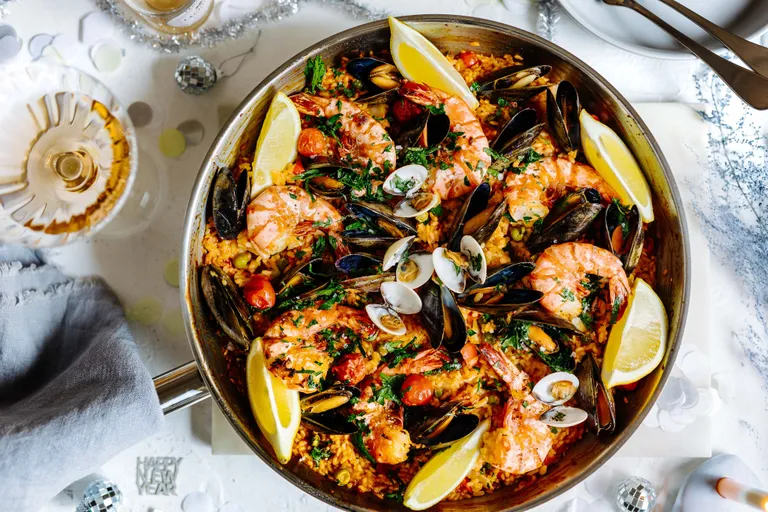 Eine Paella mit Meeresfrüchten, Garnelen, Muscheln, Venusmuscheln, Tomaten und Zitronenschnitzen in einer großen Pfanne auf dem Tisch, daneben ein Weinglas und eine Serviette.