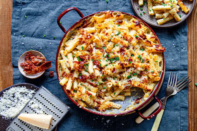 Überbackene Mac & Cheese mit geschmolzenem Käse, Speckstückchen und Schnittlauch in einer roten Pfanne, dazu geriebener Käse, Speck und Gabeln auf einem Holztisch.