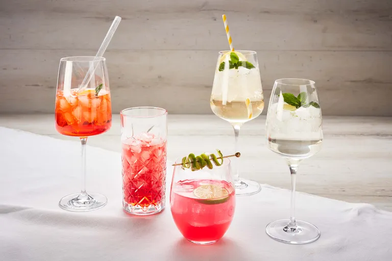 Eine Anordnung von fünf bunten Cocktails in verschiedenen Gläsern, garniert mit Minze, Zitrusfrüchten und Strohhalmen, präsentiert auf einer weißen Unterlage.
