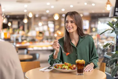 Eine Frau in einem grünen Shirt lächelt, während sie in einem Restaurant einen Salat isst; auf dem Tisch stehen ein Getränk und Besteck.