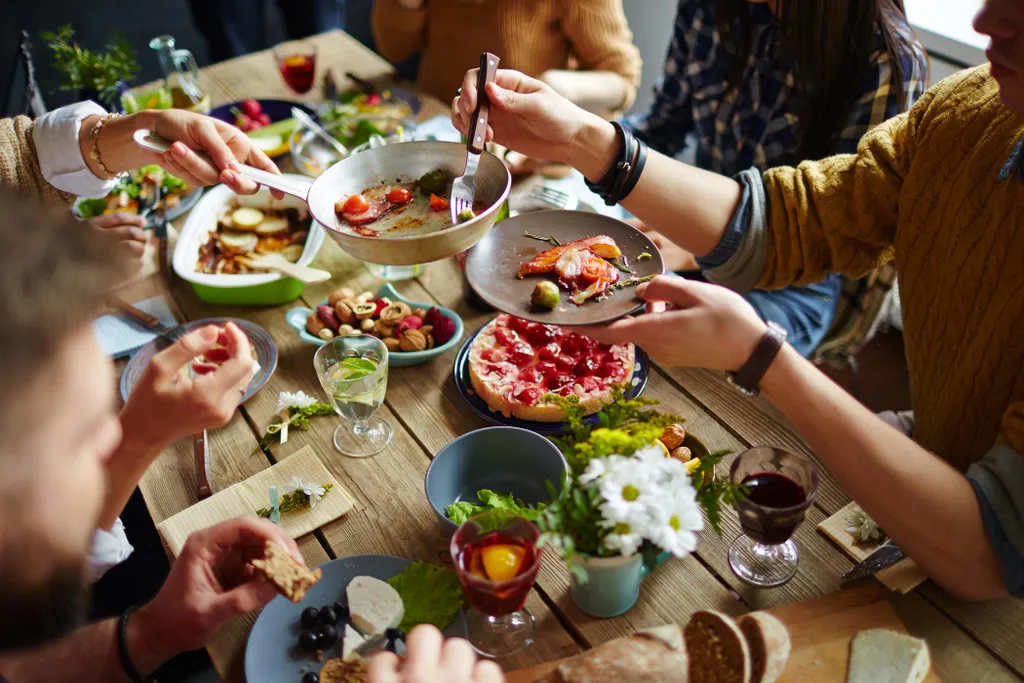 Eine Gruppe von Menschen, die gemeinsam an einem Holztisch essen; zu sehen sind Desserts, Getränke, Nüsse und farbenfrohes Geschirr mit Blumenverzierung.