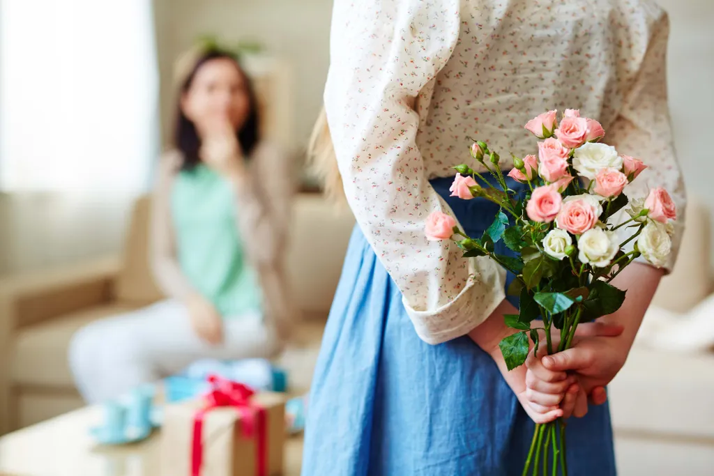 Ein Mädchen hält einen Blumenstrauß hinter dem Rücken. Im Hintergrund ist verschommen eine Frau zu erkennen
