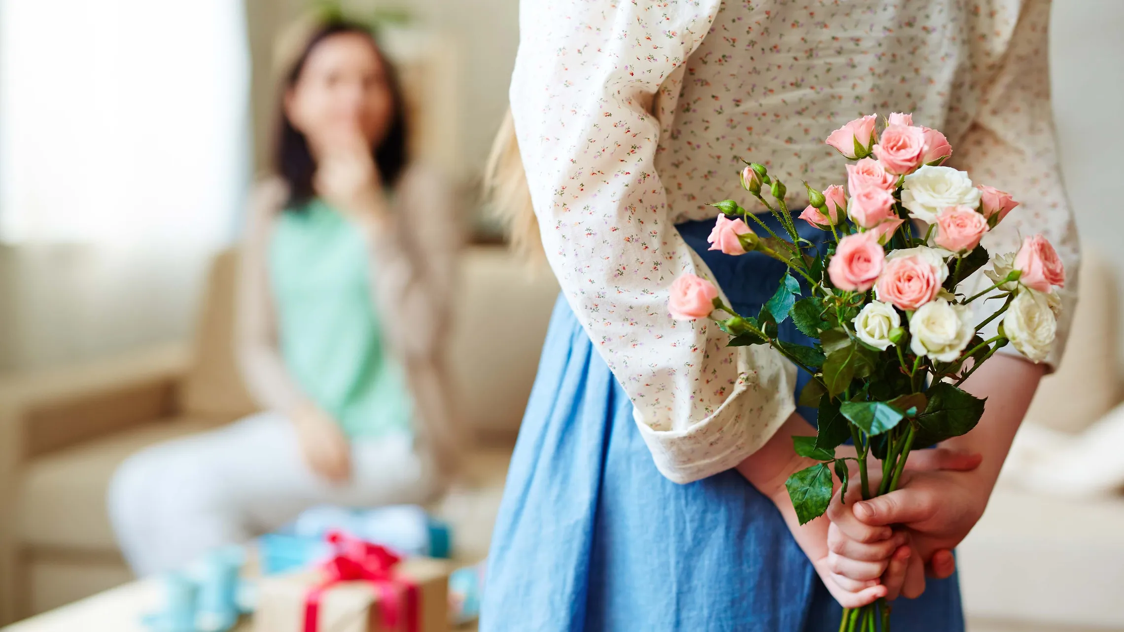 Ein Mädchen hält einen Blumenstrauß hinter dem Rücken. Im Hintergrund ist verschommen eine Frau zu erkennen