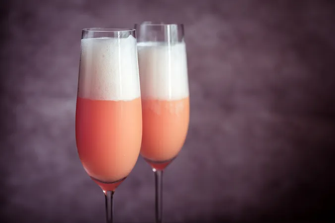 Zwei hohe Champagnergläser, gefüllt mit einem rosa-orangefarbenen Cocktail, gekrönt von schaumigem Schaum vor einem violetten Hintergrund.