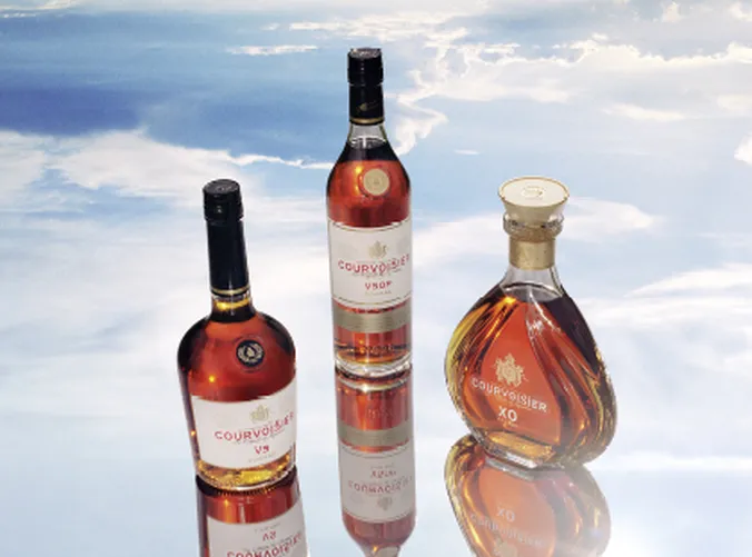 Drei Courvoisier-Cognac-Flaschen (VS, VSOP, XO) vor einem Himmel mit Wolken.