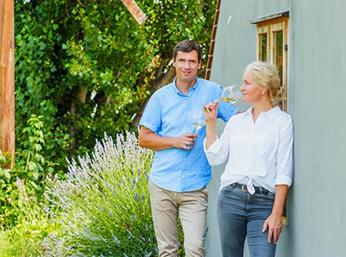 Ein Mann und eine Frau stehen im Freien, lehnen an einem Gebäude und halten Weingläser in der Hand. Sie nippt an ihrem Wein, umgeben von Grünpflanzen und Lavendel.