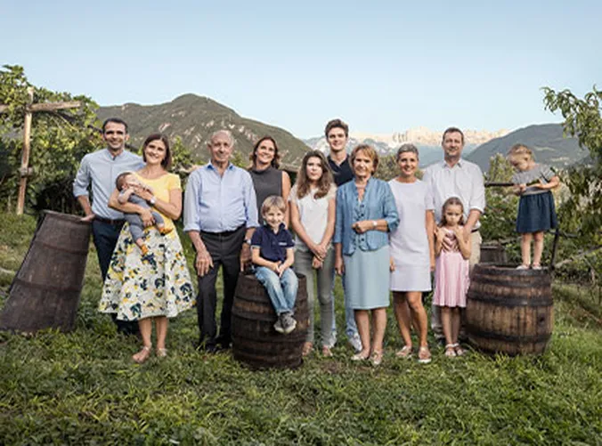 Eine Familiengruppe steht im Freien in einem Weinberg, im Hintergrund sind Berge zu sehen. Sie stehen um Holzfässer herum und bestehen aus Menschen unterschiedlichen Alters.