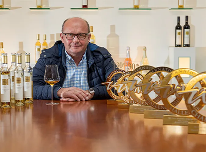 Ein Mann sitzt an einem Tisch mit Weinflaschen und mehreren Goldauszeichnungen, blickt in die Kamera und befindet sich in einem gut beleuchteten Raum, in dessen Hintergrund Weinauslagen zu sehen sind.
