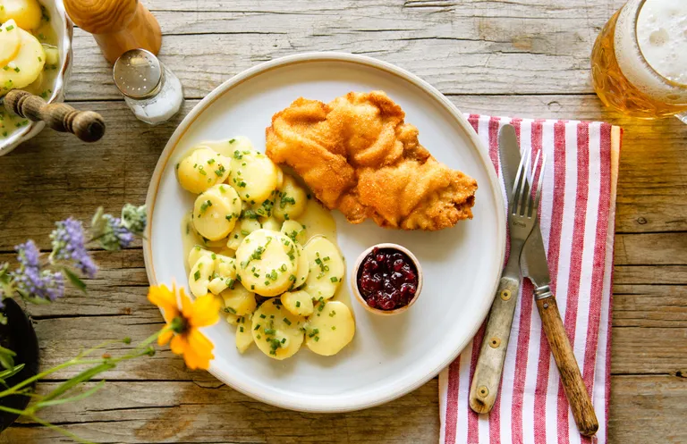 Ein Teller mit Schnitzel, Kartoffelsalat und Preiselbeersauce auf einem Holztisch, daneben eine gestreifte Serviette, Besteck und ein Bierkrug.