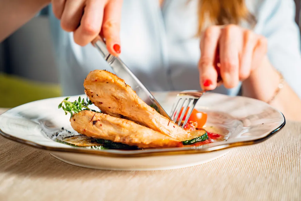 Person schneidet gegrillte Hähnchenbrust mit Zucchini und Tomaten auf einem Teller.