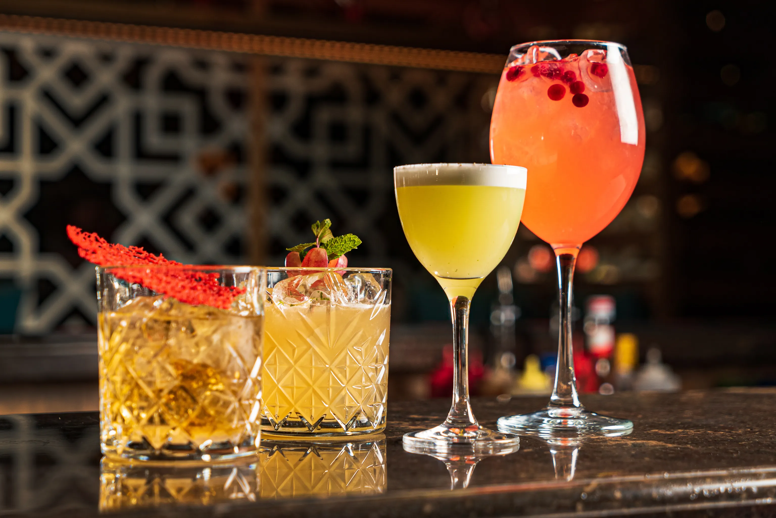 Eine Auswahl von vier farbenfrohen Cocktails auf einer Bartheke, jedes in einem anderen Glas und mit einzigartigen Garnituren.