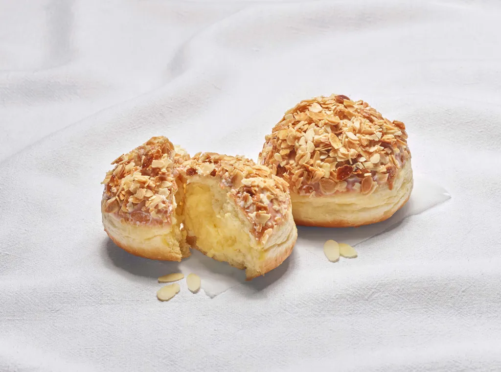 Zwei Bienenstich-Krapfen aus der INTERSPAR-Backstube legen auf einem weißen Tuch wobei einer der Krapfen mittig halbiert ist