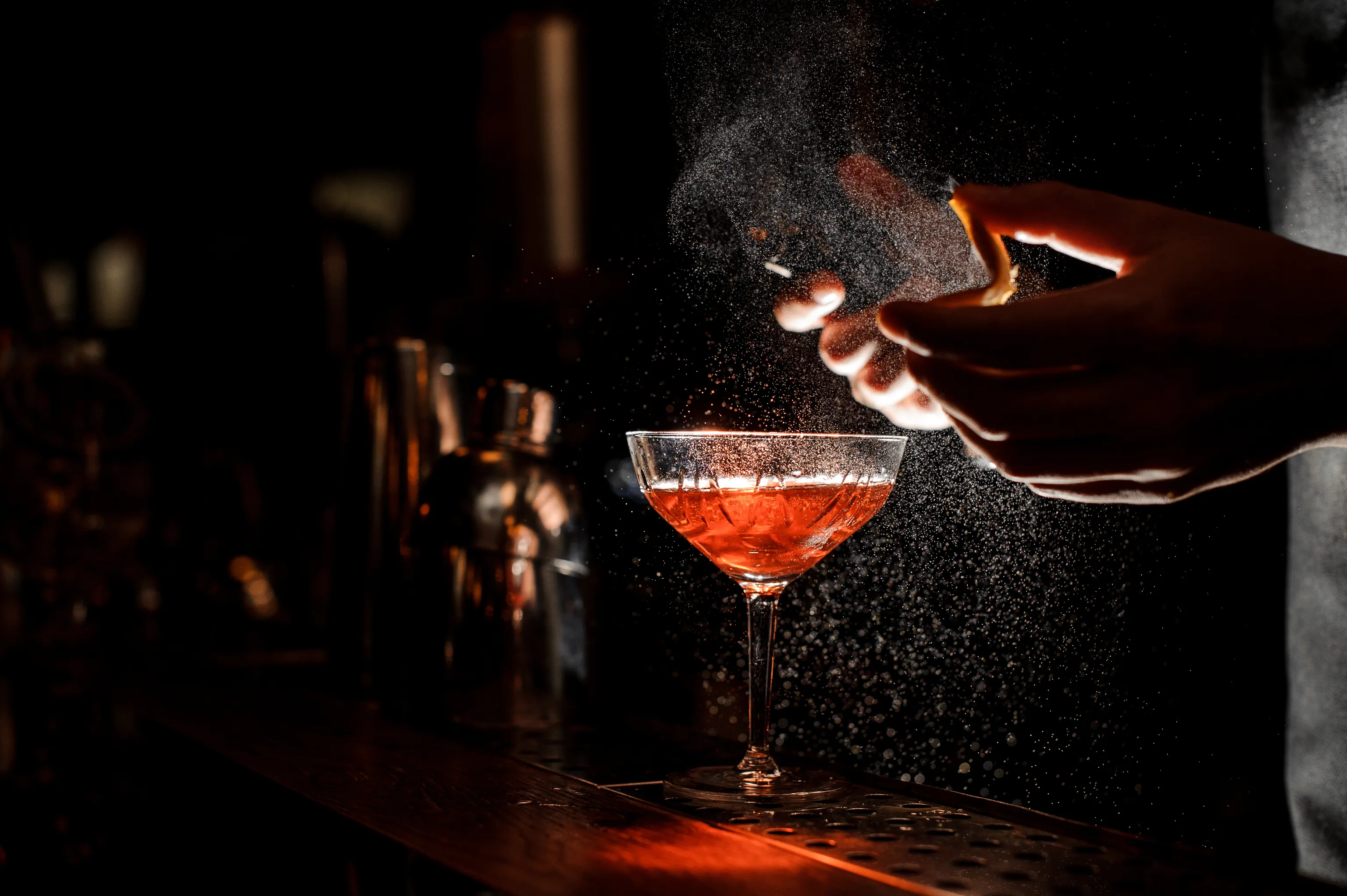 Ein Barkeeper, der einen Cocktail mit einer Orangenspirale garniert, wobei ein aromatischer Nebel aufsteigt, bei gedämpftem Licht und mit einem Shaker im Hintergrund.