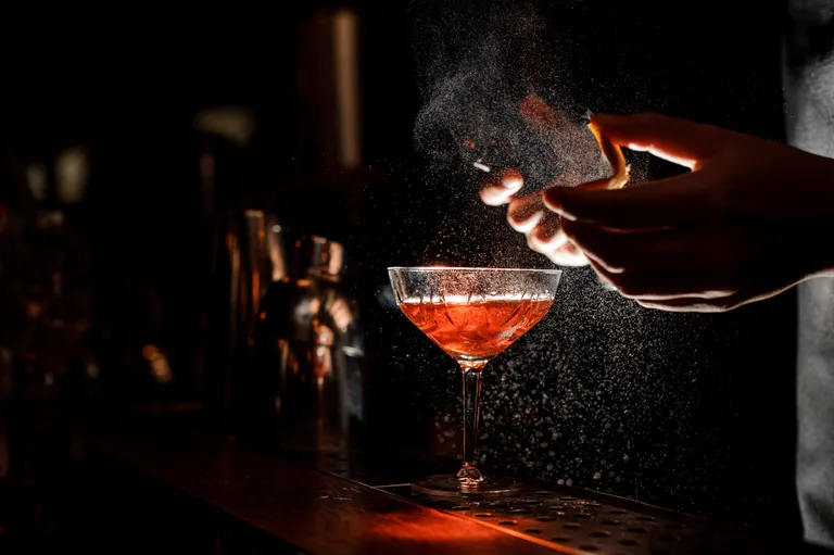 Ein Barkeeper, der einen Cocktail mit einer Orangenspirale garniert, wobei ein aromatischer Nebel aufsteigt, bei gedämpftem Licht und mit einem Shaker im Hintergrund.