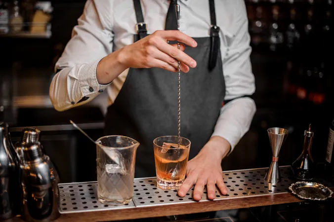 Ein Barkeeper, der mit einem Metalllöffel einen Cocktail umrührt; er trägt ein weißes Hemd und eine dunkle Schürze. Auf der Theke sind Barutensilien zu sehen.