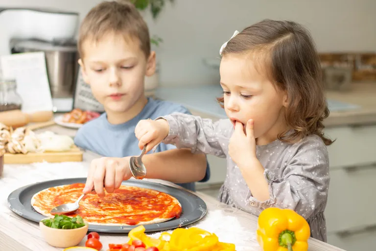 Zwei Kinder bestreichen eine Pizza mit Tomatensauce