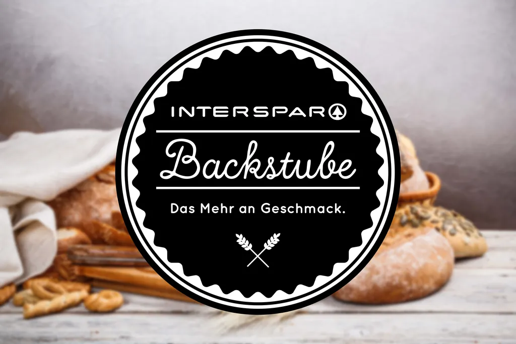 Schwarzes, rundes Logo mit weißen Akzenten und der Aufschrift "INTERSPAR Backstube. Das Mehr an Geschmack." auf einem unscharfen Hintergrund auf dem Brot und Gebäck zu erkennen ist