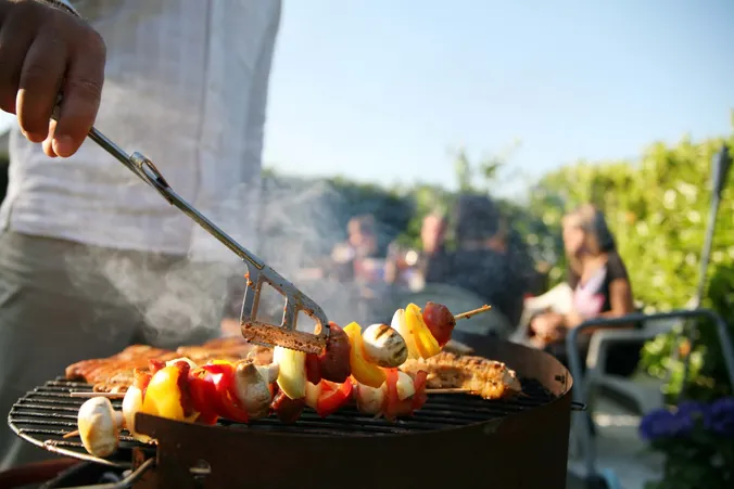 Eine Person beim Grillen eines Spießes mit Gemüse auf einem Grill im Sonnenschein