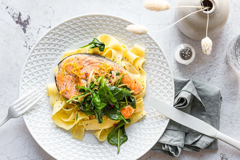 Ein Teller mit Pappardelle, garniert mit gegrilltem Lachs, Spinat und Zitronenschale, serviert auf einem strukturierten Teller, daneben Besteck und eine dekorative Vase.