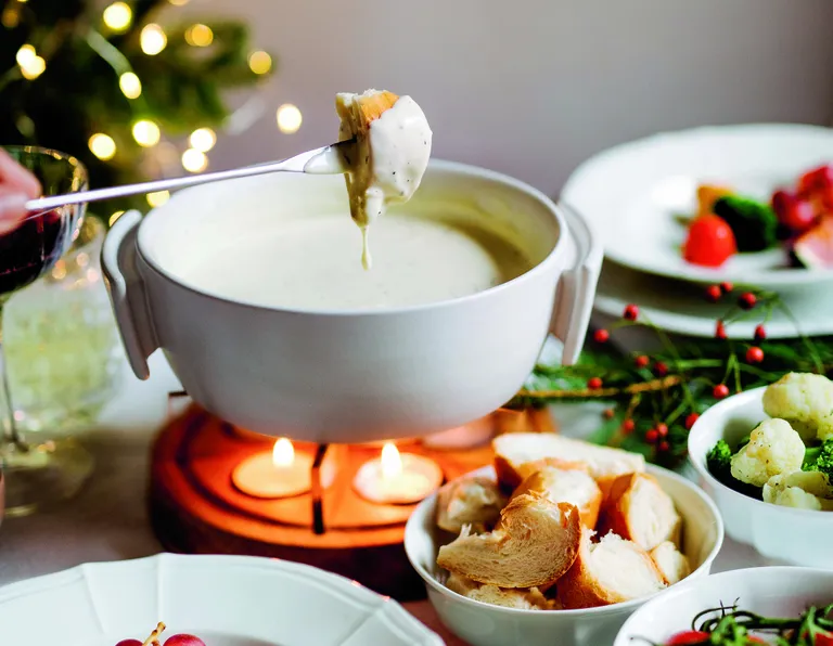 Eine festliche Fondue-Dekoration mit Brotwürfeln, die in cremigen Käse getaucht werden, umgeben von Gemüse und beleuchtet von warmem Kerzenlicht.