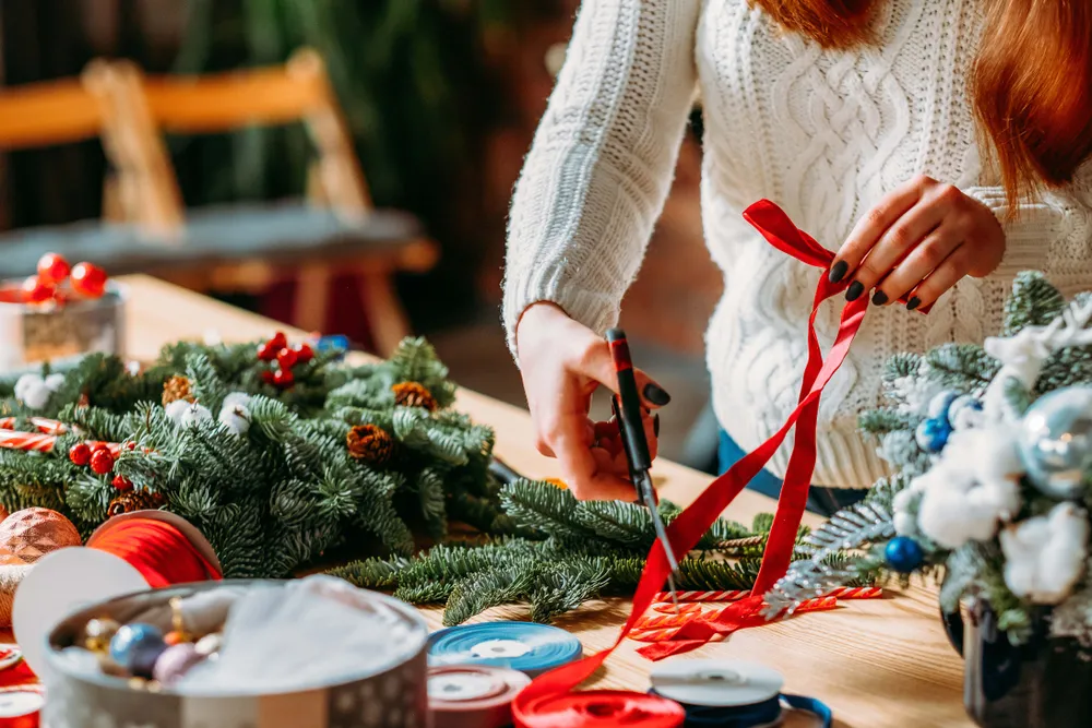 Eine Person in einem weißen Pullover schmückt einen Weihnachtskranz mit einem roten Band auf einem Tisch, der mit festlichen Ornamenten und Materialien bedeckt ist.