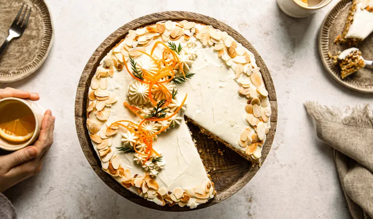 Karottenkuchen mit Frischkäse-Glasur, Mandelscheiben und dekorativen Karottenspiralen auf einem Holzständer, dazu Kaffee und ein Stück Kuchen auf einem Teller daneben.