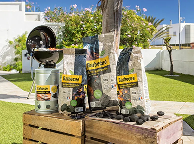 Eine Aufstellung von diversen Grill-Artikeln der Eigenmarken Barbecue im Garten. Im Hintergrund sieht man einen Kugelgrill stehen mit Lebensmitteln, die gerade gegrillt werden.