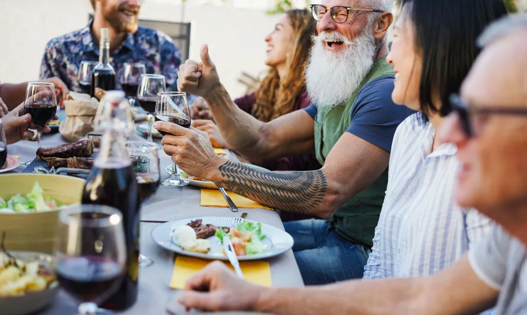 Eine fröhliche Gruppe von Menschen, die im Freien ein Essen genießen, Weingläser in der Hand halten und gemeinsam am Tisch mit Salaten und Tellern voller Speisen lachen.