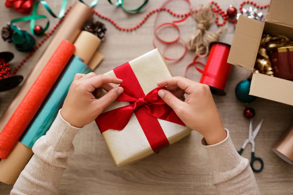 Hände, die ein rotes Band an einer Geschenkbox befestigen, umgeben von Rollen Geschenkpapier, Bändern und einer Schere auf einem Basteltisch.