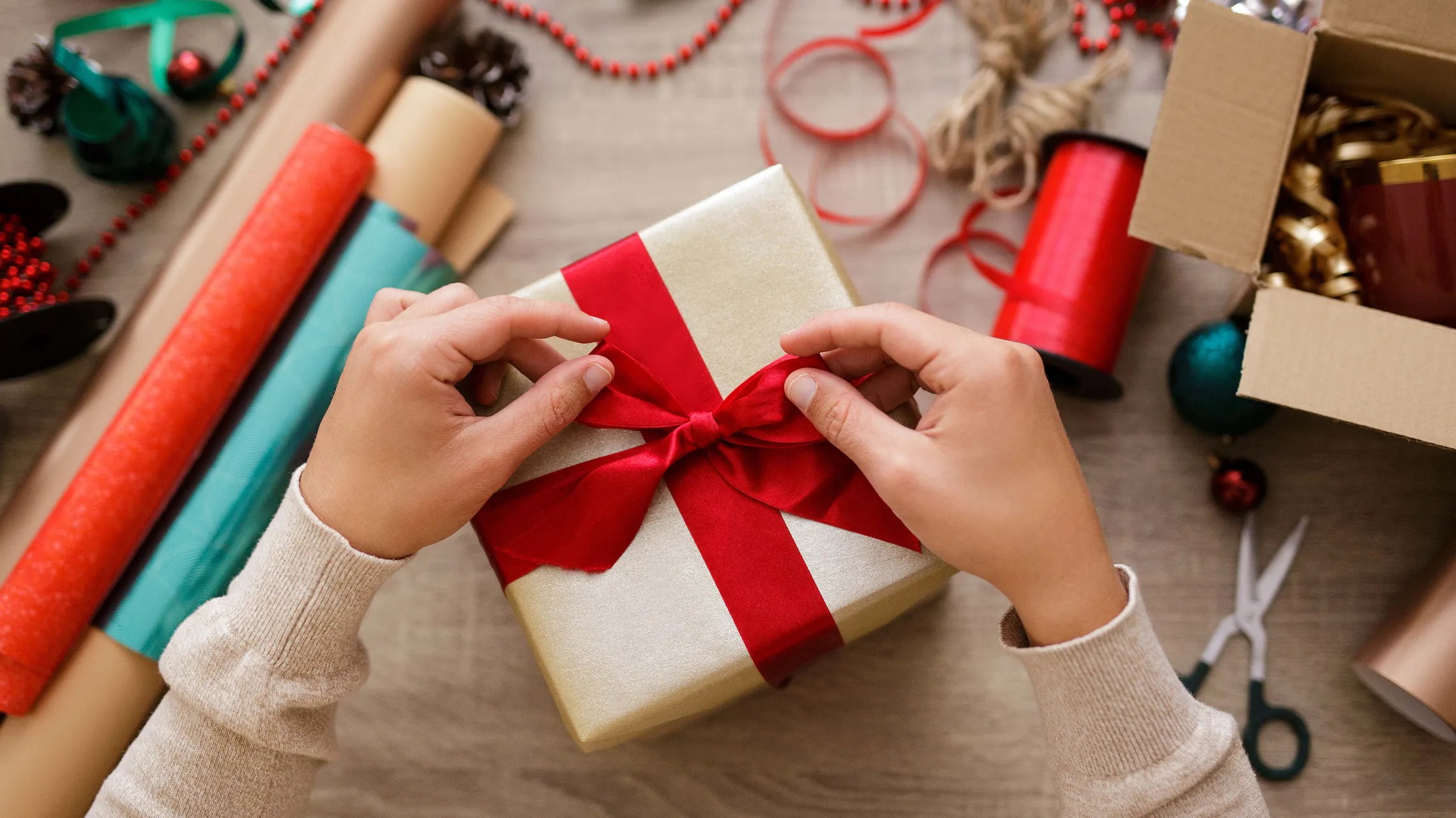 Hände, die ein rotes Band an einer Geschenkbox befestigen, umgeben von Rollen Geschenkpapier, Bändern und einer Schere auf einem Basteltisch.