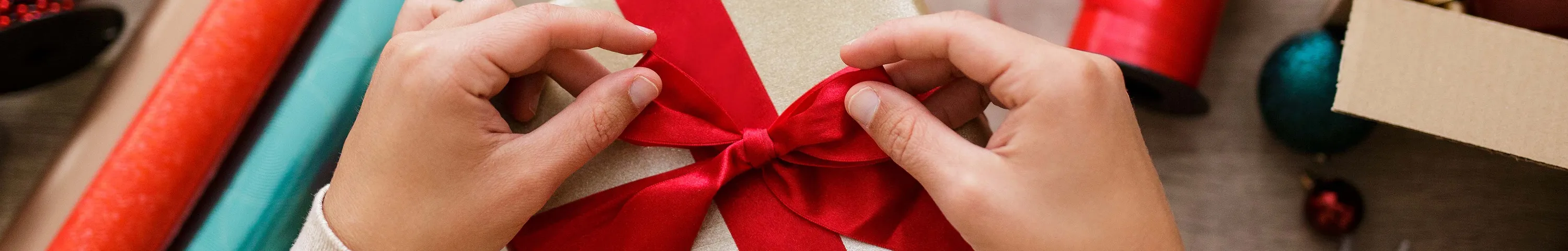 Hände, die ein rotes Band an einer Geschenkbox befestigen, umgeben von Rollen Geschenkpapier, Bändern und einer Schere auf einem Basteltisch.