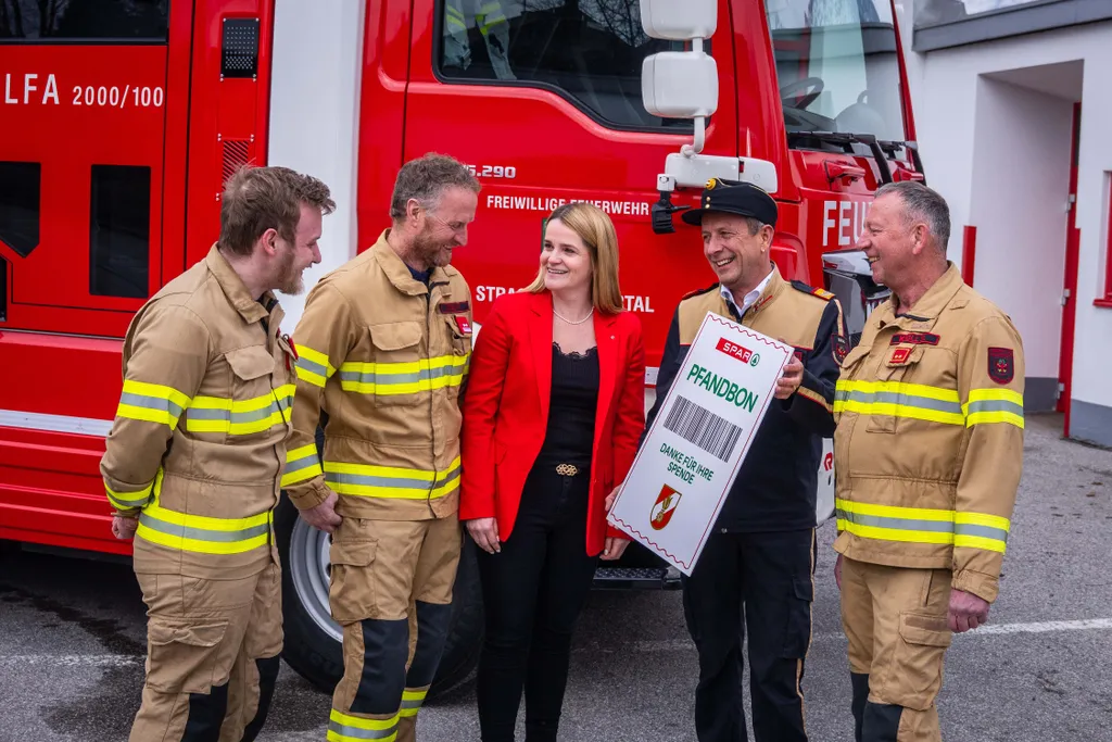 Eine junge Frau in einem roten Blazer und mehrere lachende Männer in Feuerwehruniformen mit einem großen Pfandbon in den Händen