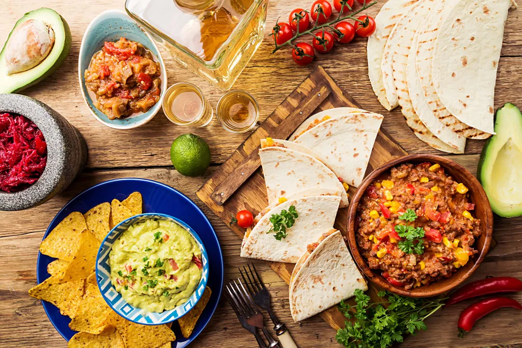 Ein mexikanisches Festmahl mit Tacos, Salsa, Guacamole, Nachos, Avocado, Kirschtomaten, Limette, Rote-Bete-Dip, Tequila und Tortillas auf einem Holztisch.