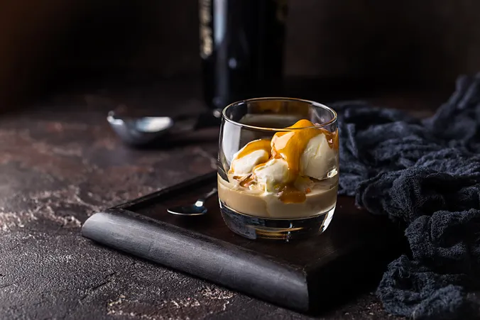 Ein Glas Cremelikör mit Vanilleeis und Karamellsauce auf einem dunklen Holztablett, im Hintergrund ein Löffel.