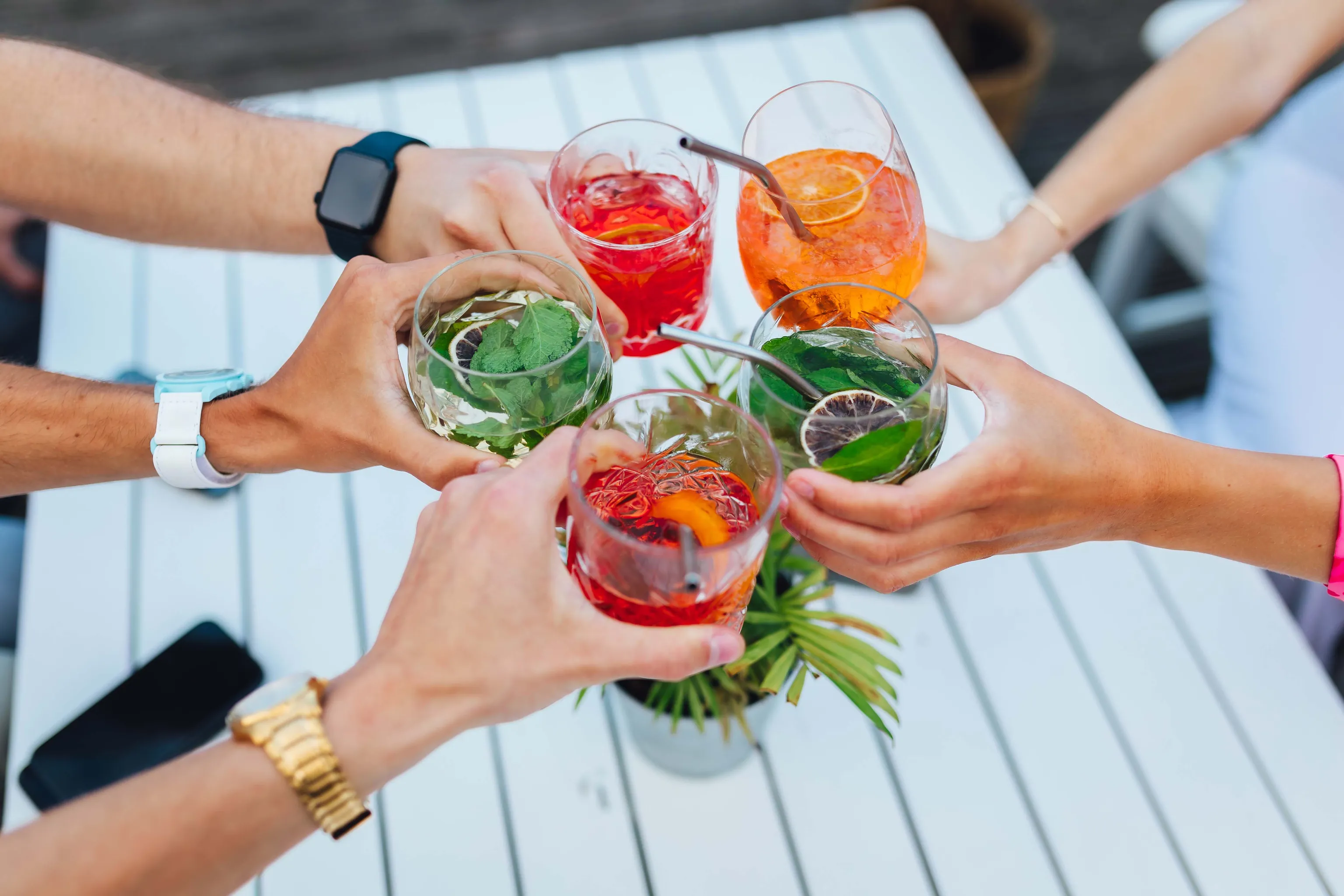 Fünf Menschen stoßen mit bunten Cocktails und Garnituren an einem weißen Tisch an und feiern gemeinsam.