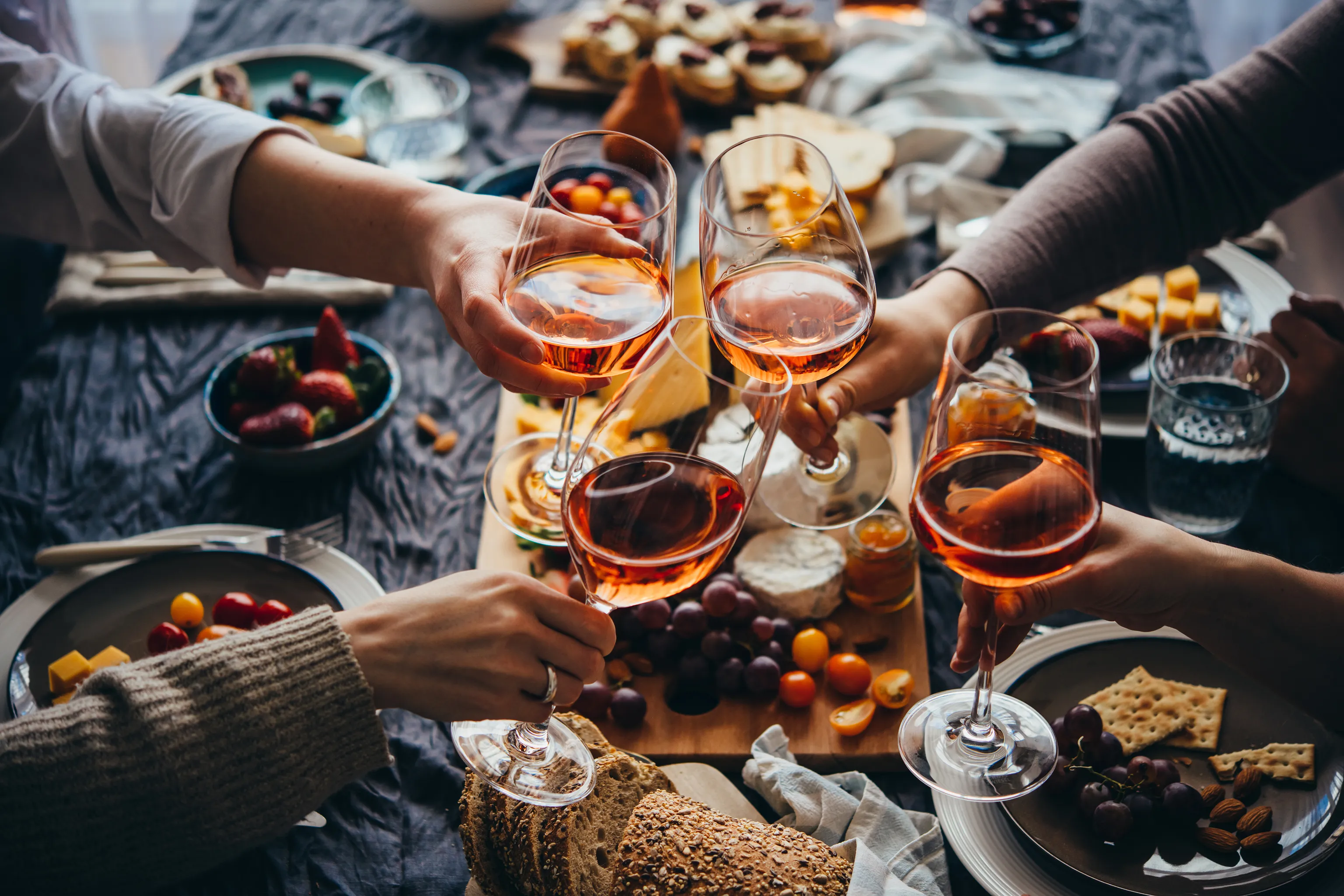 Vier Hände stoßen mit Gläsern Roséwein an einem Tisch an, der mit Obst, Käse, Brot und Crackern gedeckt ist, und sorgen so für eine festliche Atmosphäre.