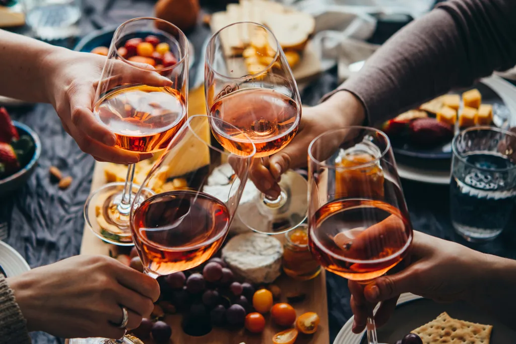 Hände, die mit Gläsern Roséwein auf eine Wurst- und Käseplatte mit Käse, Weintrauben und Tomaten anstoßen.