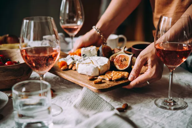 Hände, die ein Brettchen mit verschiedenen Käsesorten und Feigen auf einen gedeckten Tisch mit Roséwein-Gläsern stellen