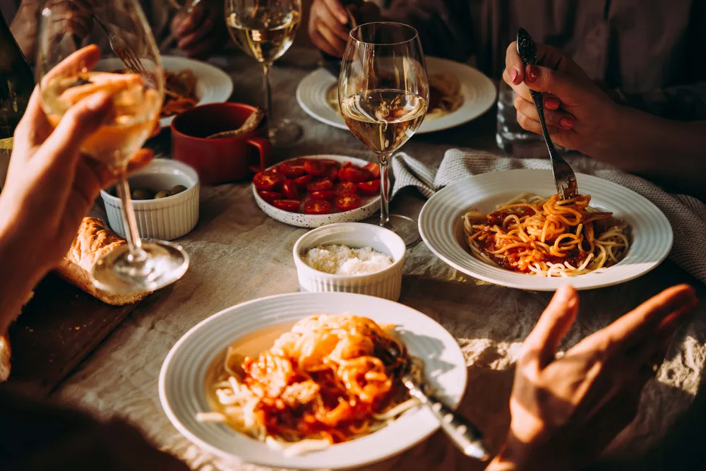 Ein gedeckter Tisch mit Spaghetti, Weingläsern, Kirschtomaten und Brot. Die Gäste sitzen beim Essen und genießen gemeinsam die Mahlzeit in warmer Beleuchtung.