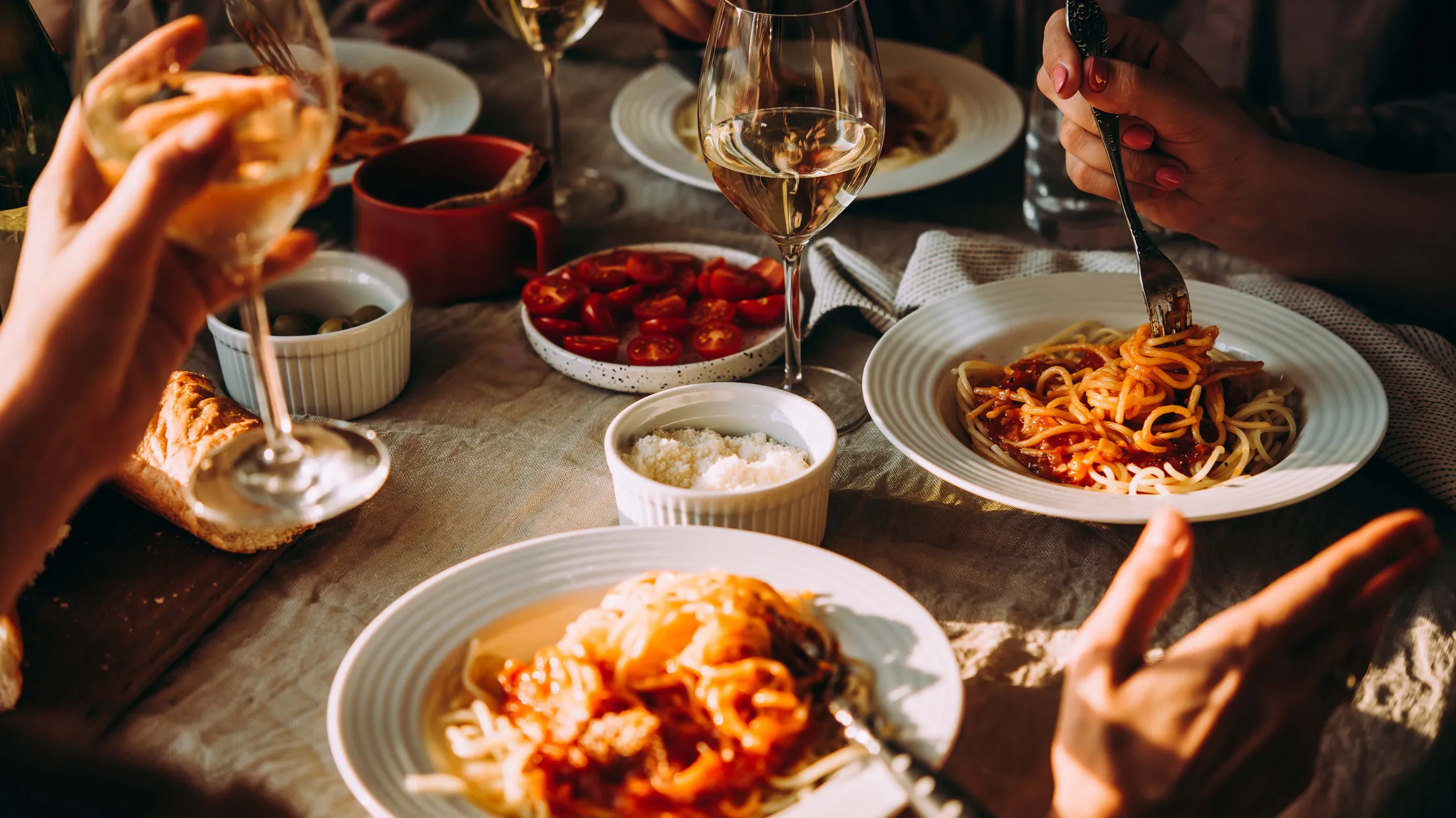 Ein gedeckter Tisch mit Spaghetti, Weingläsern, Kirschtomaten und Brot. Die Gäste sitzen beim Essen und genießen gemeinsam die Mahlzeit in warmer Beleuchtung.