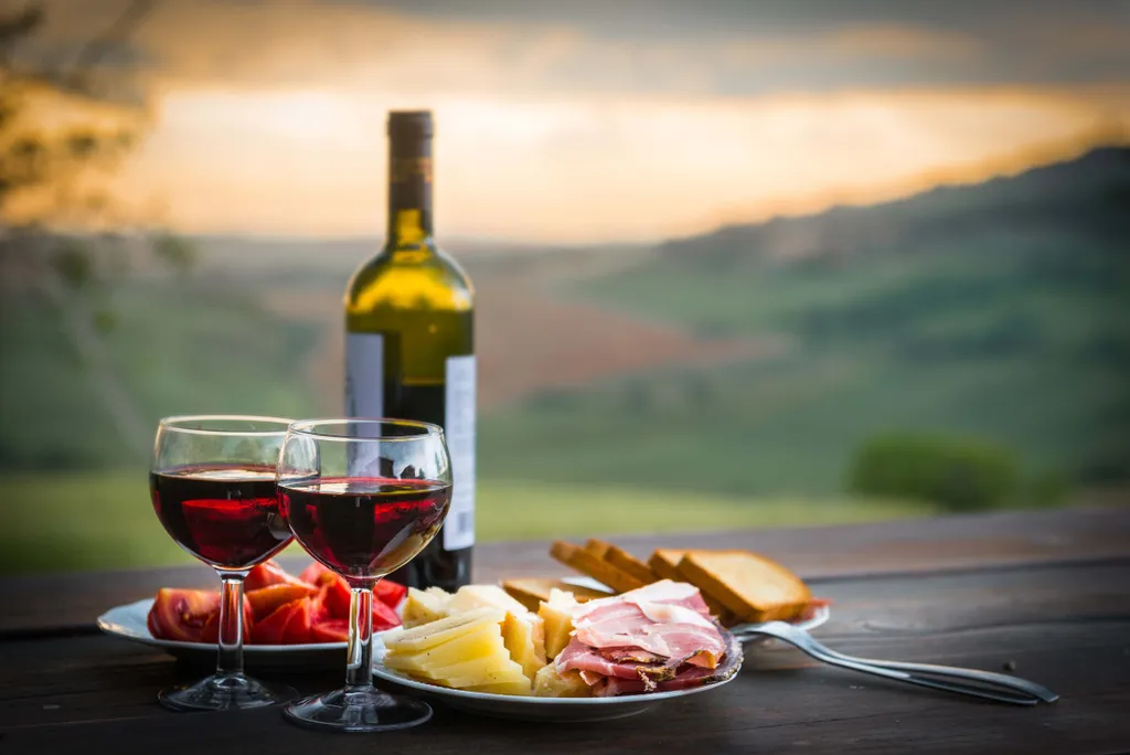 Zwei Gläser mit Rotwein und eine Rotweinflasche neben einem Teller mit Schinken- und Käseaufschnitt, Brot und Tomaten