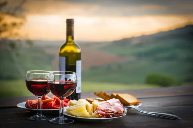 Zwei Gläser mit Rotwein und eine Rotweinflasche neben einem Teller mit Schinken- und Käseaufschnitt, Brot und Tomaten