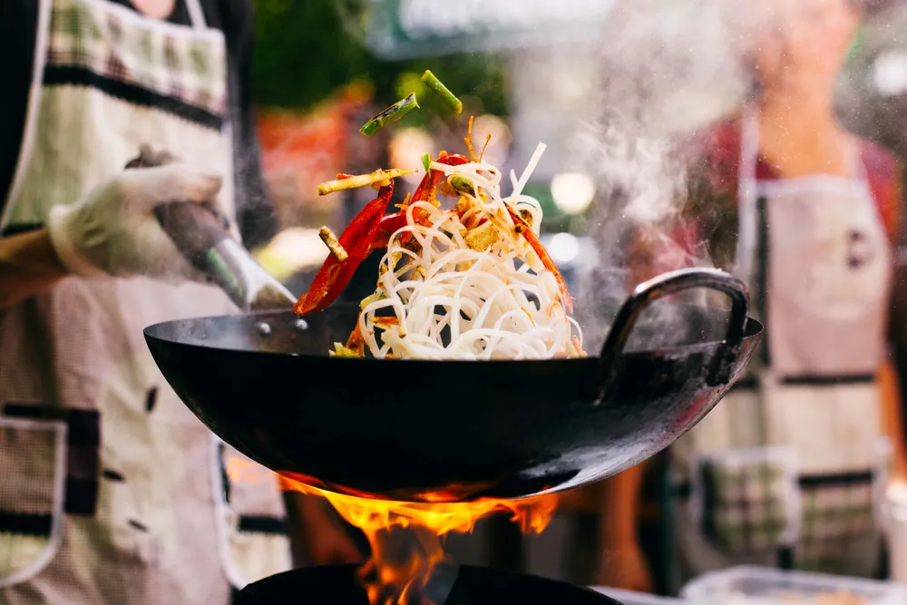 Ein Koch, der Nudeln und Gemüse in einem flammenden Wok schwenkt, während Rauch aufsteigt; er trägt eine Schürze und einen Handschuh.