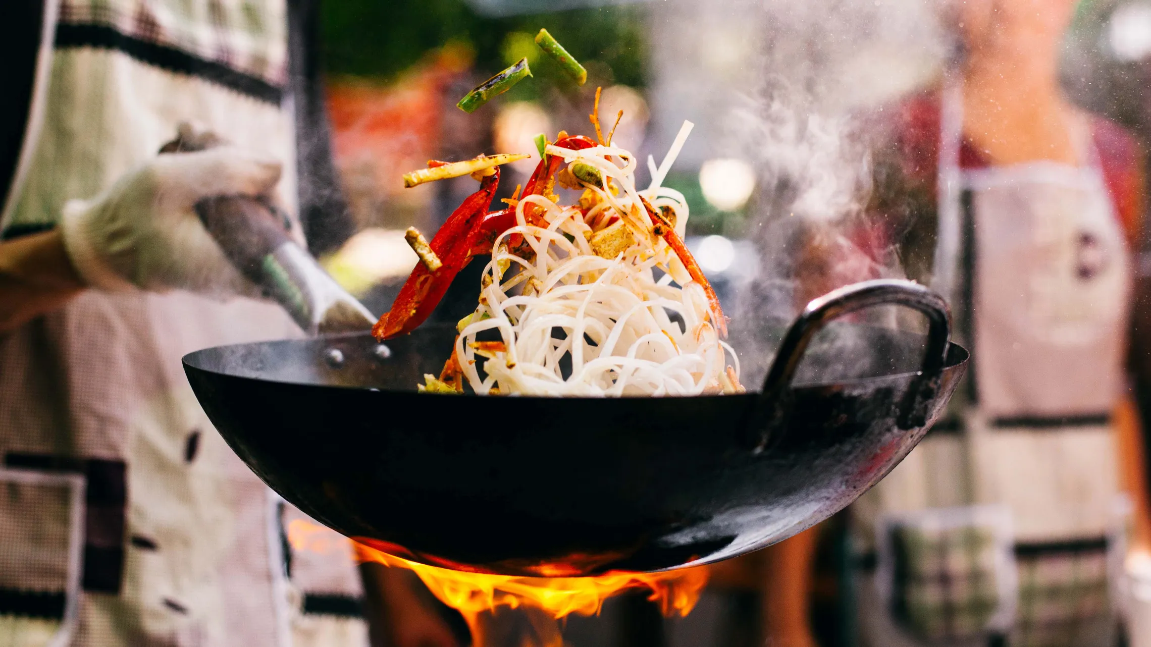 Ein Koch, der Nudeln und Gemüse in einem flammenden Wok schwenkt, während Rauch aufsteigt; er trägt eine Schürze und einen Handschuh.